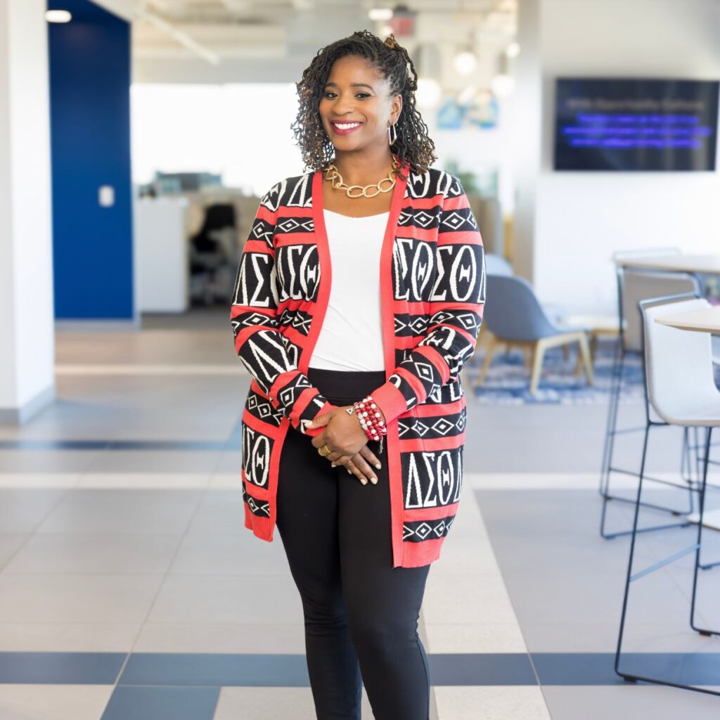 Black woman with dreads, standing, smiling, hands folded in front wearing a white crew neck shirt and a thigh length delta Sigma theta sweater