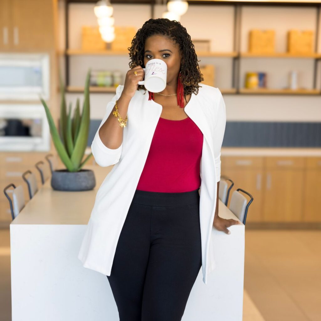 Black woman, DST, wearing red leather earrings, with white blazer and red crew neck shirt underneath. She is joyful, happy, and smiling Drinking coffee in a minimalist kitchen